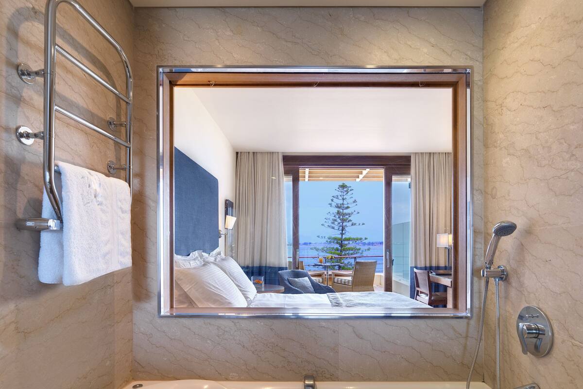 Un baño moderno con un espejo grande, una bañera, una ducha y vistas a un balcón con comedor y un árbol.