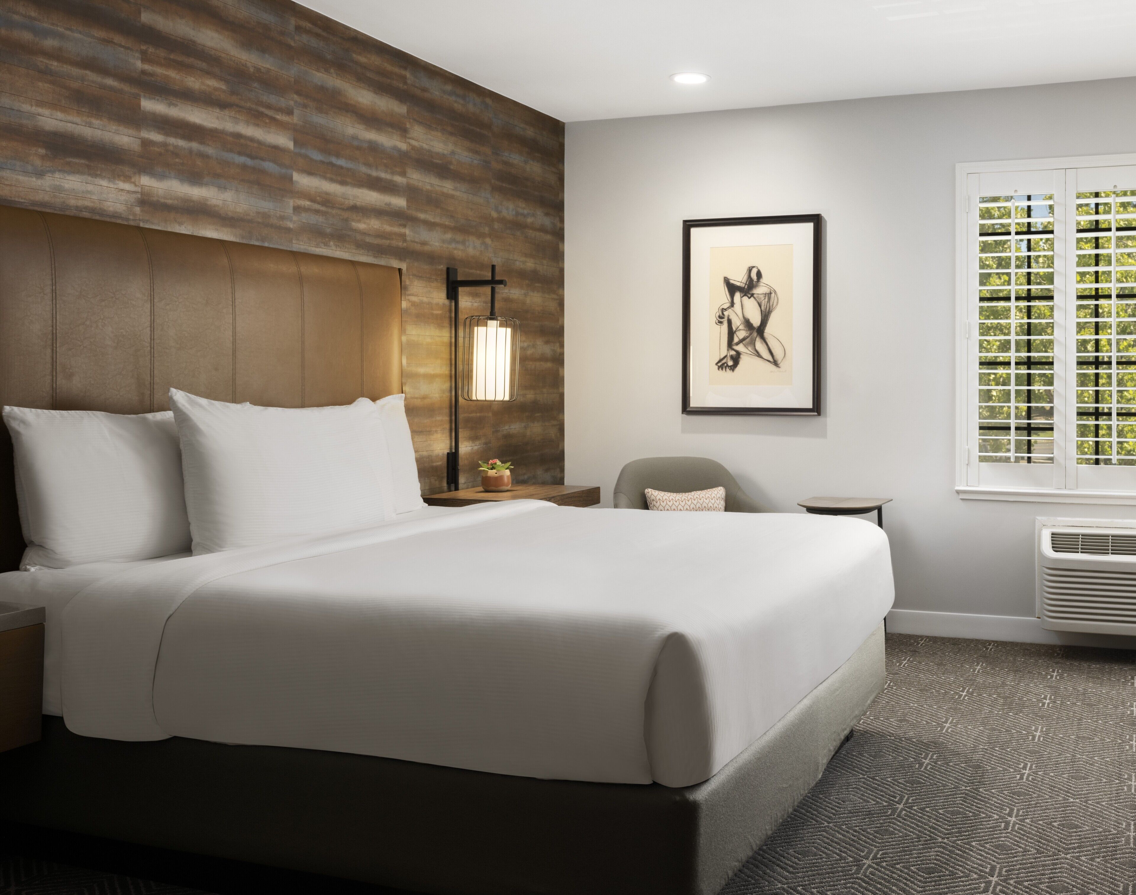 A hotel room with a large bed, a nightstand, a chair, a framed artwork, a window with blinds, and a wall-mounted heater.
