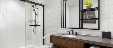 A modern bathroom with a glass shower enclosure, a white toilet, a wooden vanity with a sink, and a mirror with a framed artwork.