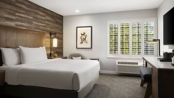 A hotel room with a large bed, a desk, a chair, a window with blinds, a framed picture on the wall, and a wall-mounted heater.