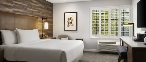 A hotel room with a large bed, a desk, a chair, a window with blinds, a framed picture on the wall, and a wall-mounted heater.