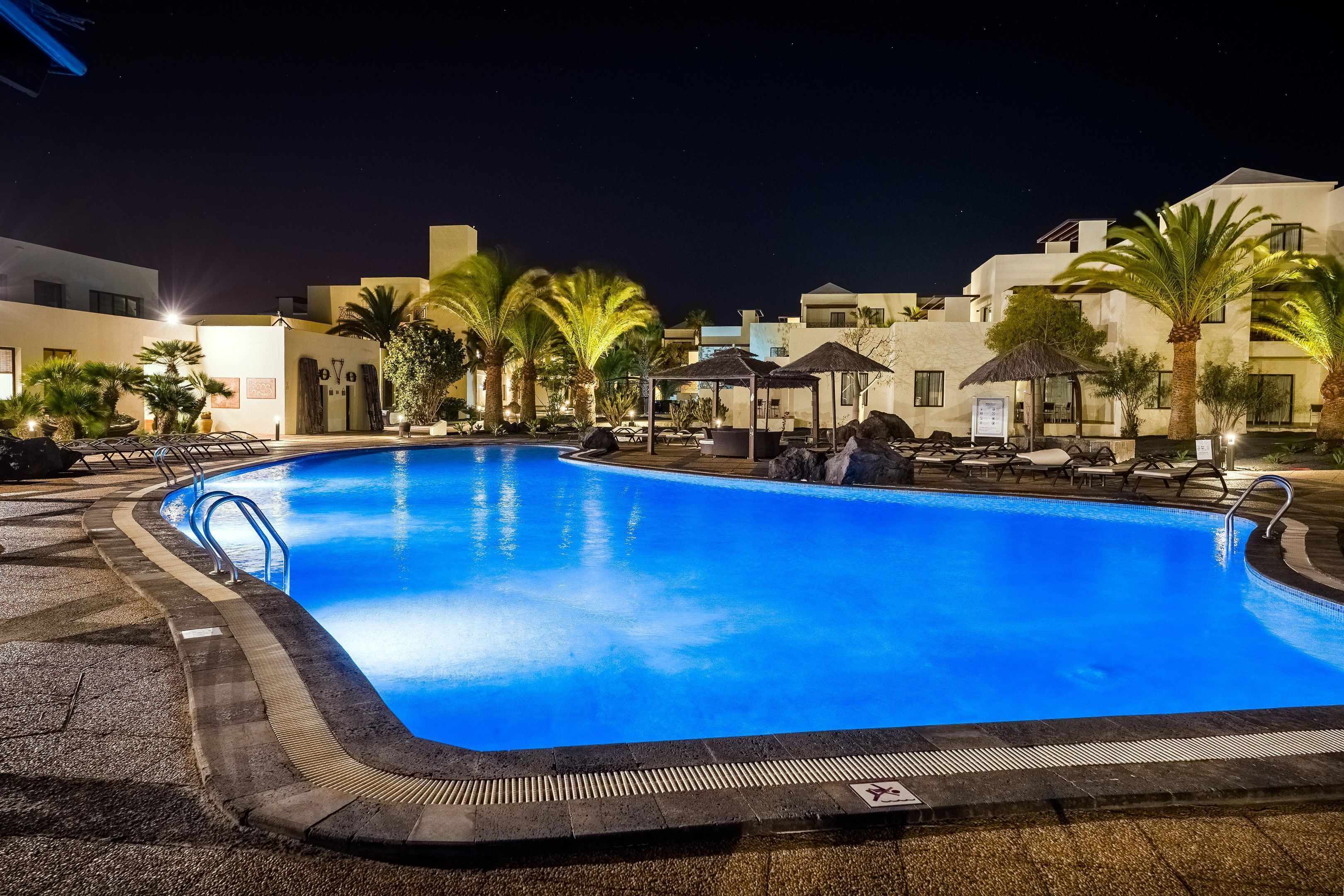 A nighttime swimming pool with lounge chairs and palm trees, surrounded by buildings with warm lighting.