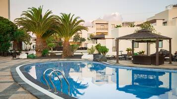 Una piscina con sillones de descanso y un pabellón, rodeada de palmeras y un edificio con techo plano.
