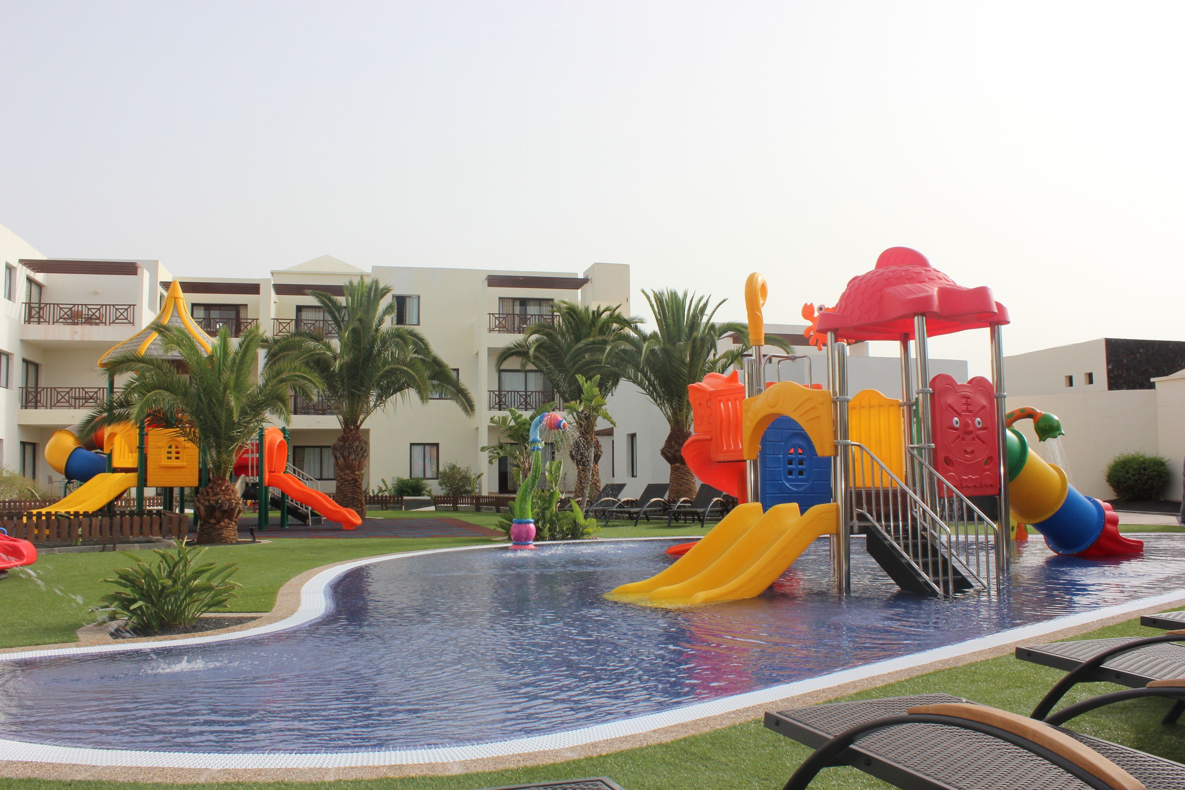 A pool area with slides and water features, surrounded by palm trees and a building with balconies.