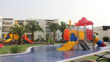 A pool area with slides and water features, surrounded by palm trees and a building with balconies.