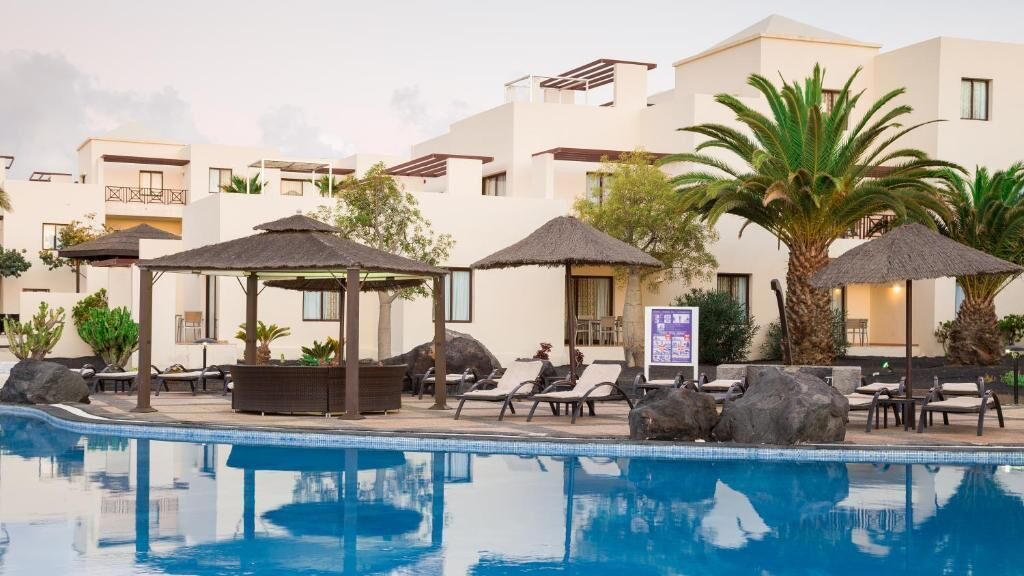 Zona de piscina con sillones de descanso, sombrillas y un edificio con balcones.
