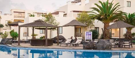 Zona de piscina con sillones de descanso, sombrillas y un edificio con balcones.