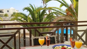 A table set with a fruit platter, drinks, and a cup on a balcony overlooking a pool and palm trees. - Vitalclass Lanzarote Resort (Teguise)