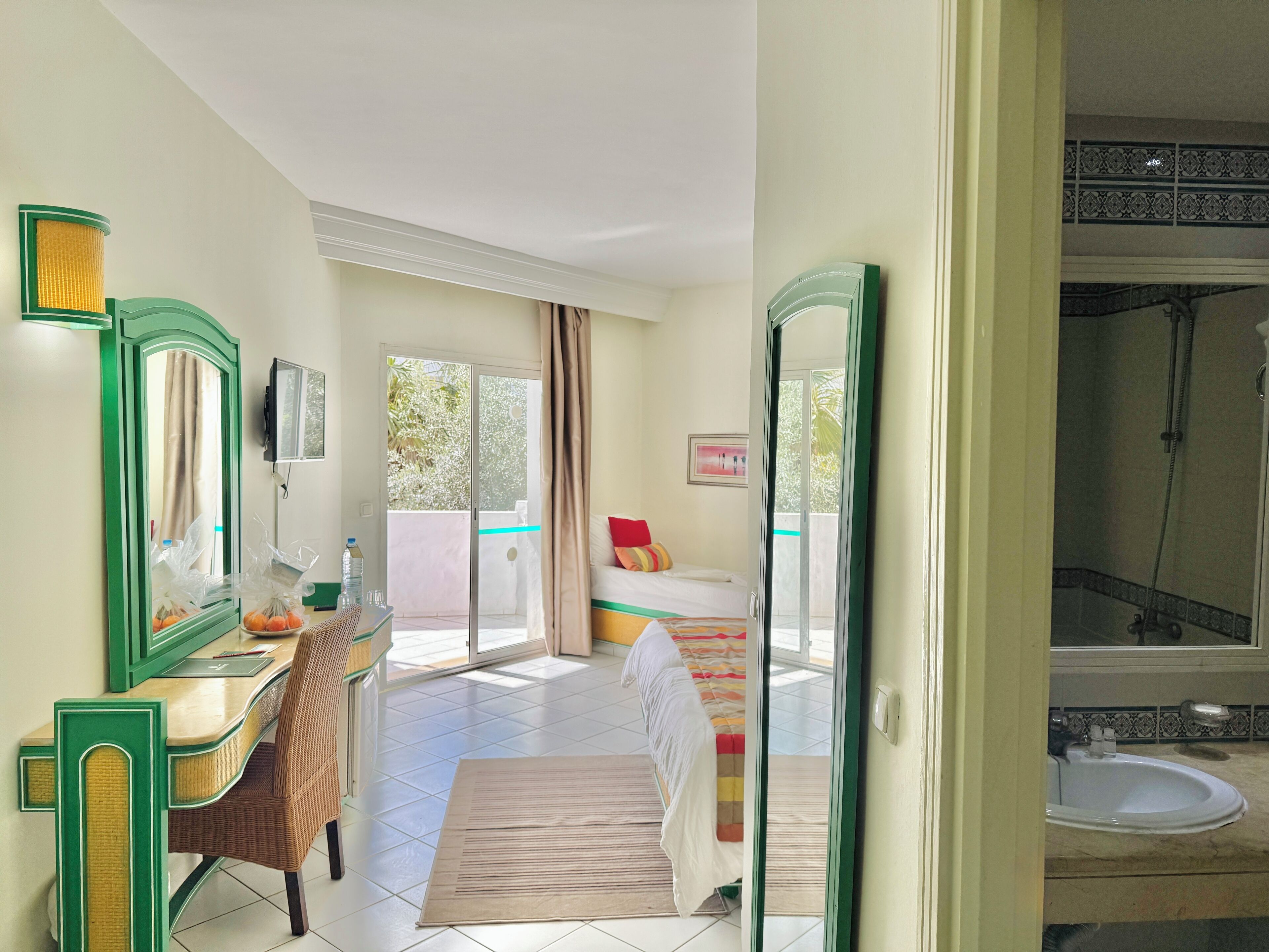 Ein Hotelzimmer mit Bett, Schreibtisch mit Stuhl, Bad mit Waschbecken und Dusche sowie ein Balkon mit Blick ins Grüne.