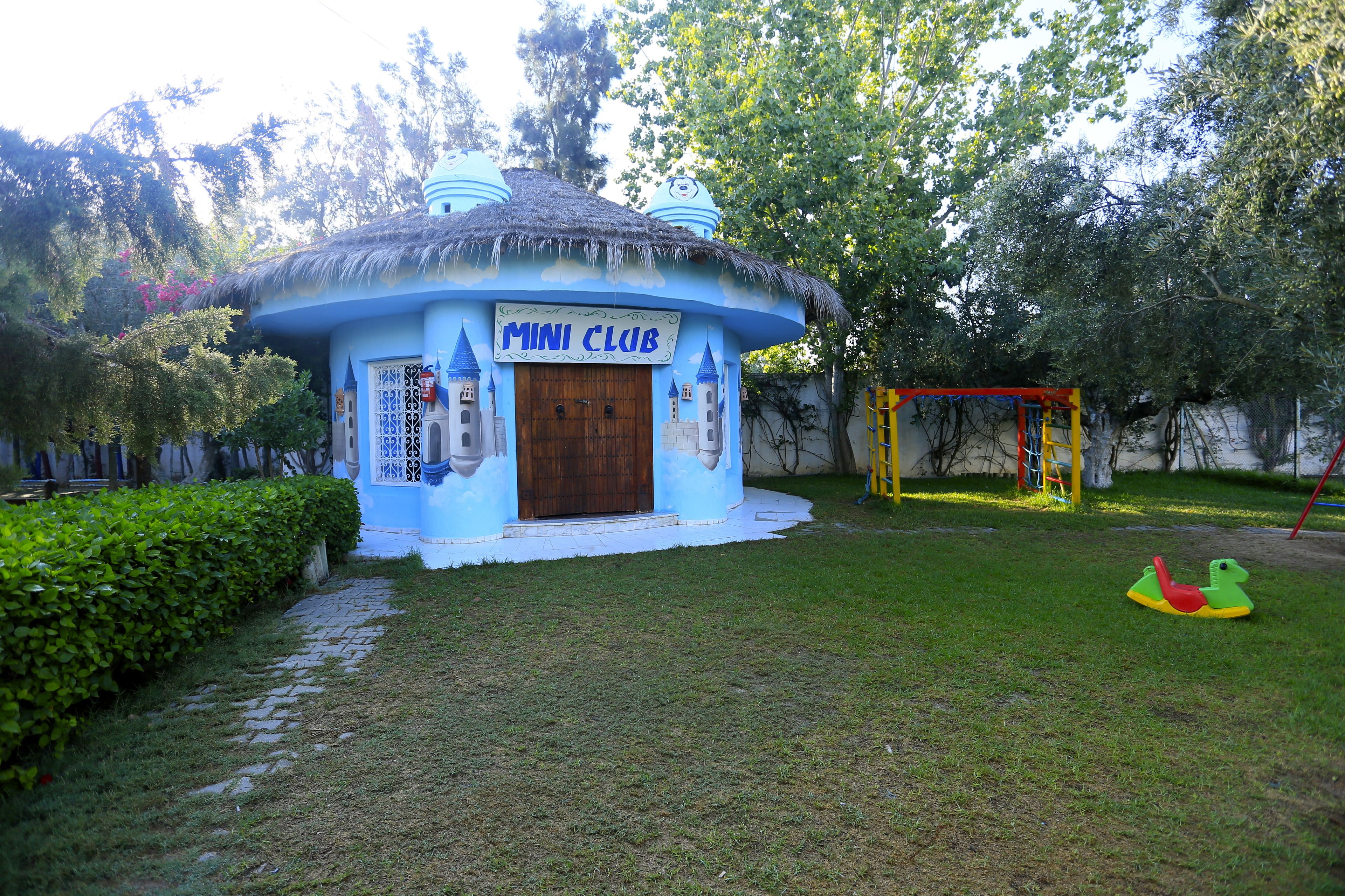 A blue building labeled "MINI CLUB" with a thatched roof, surrounded by a grassy area and trees.