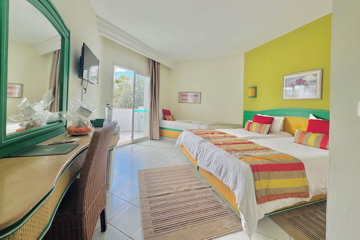 Une chambre d’hôtel avec deux lits, un grand miroir, un bureau avec une chaise et une vue sur un espace extérieur accessible par une porte coulissante.