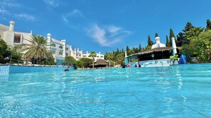 Ein großer Pool mit klarem, blauem Wasser, umgeben von Palmen und einem Gebäude mit heller Fassade und mehreren Türmen.