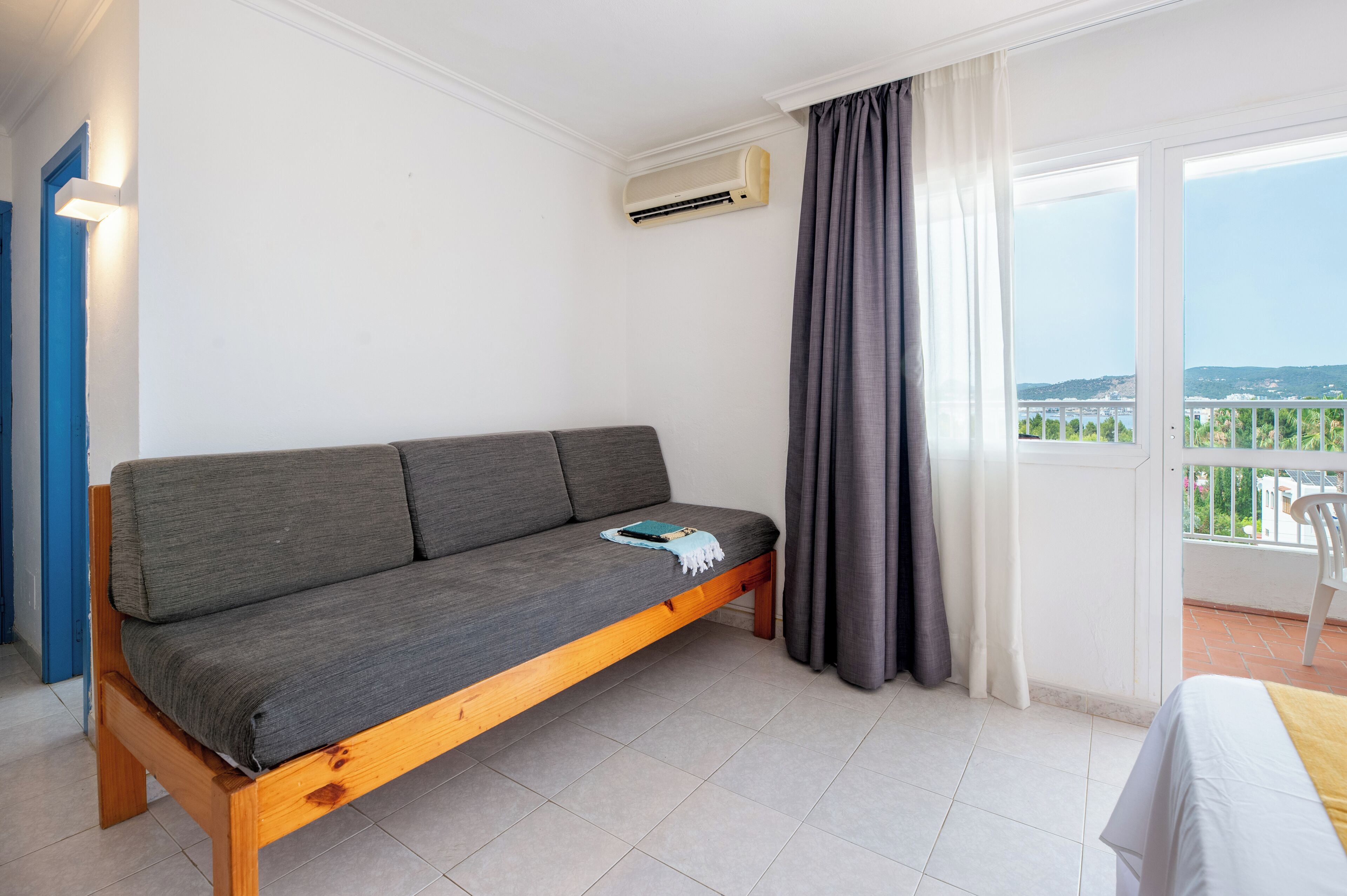 A room with a grey sofa, a small table with a book, a balcony with a view, and an air conditioning unit.