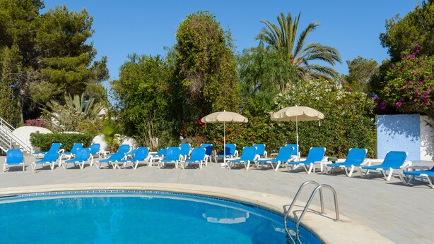 Ein Poolbereich mit blauen Liegestühlen und Sonnenschirmen, umgeben von Bäumen und blauem Himmel.