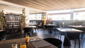 Un espace repas moderne avec un plancher en bois, des chaises noires et des tables préparées pour le petit-déjeuner. On y trouve également des plantes en pot et une vitrine en verre contenant des couverts.