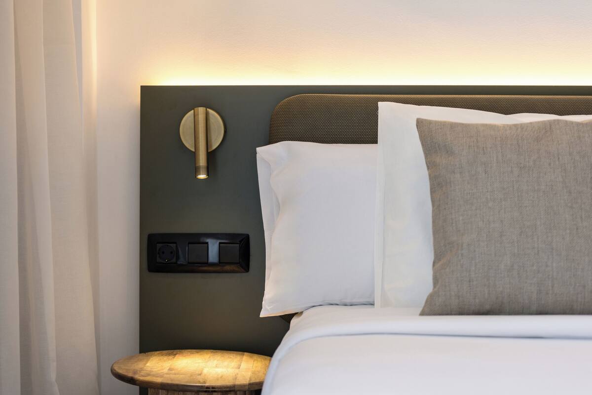Un lit bien fait, avec des oreillers blancs et gris, un luminaire rond fixé au mur et une prise de courant sur le mur.
