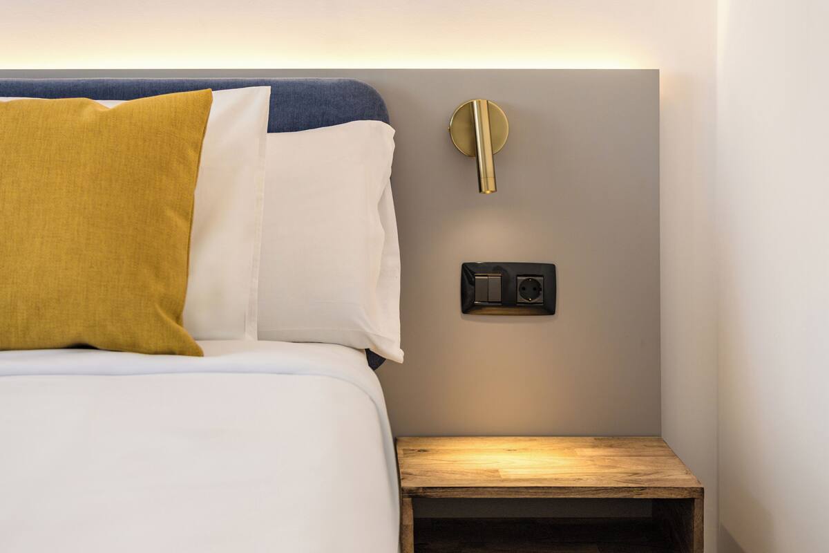 Un lit bien fait, avec une tête de lit bleue, un oreiller de couleur moutarde et un oreiller blanc. On y trouve une petite table de chevet en bois, avec une lumière en dessous, ainsi qu’un interrupteur ou une prise fixée au mur.