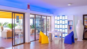 A modern lobby with glass doors leading to an outdoor seating area, a white table with chairs, and a bookshelf. - Paraiso del Sol & Paradero Apartamentos (Adeje)