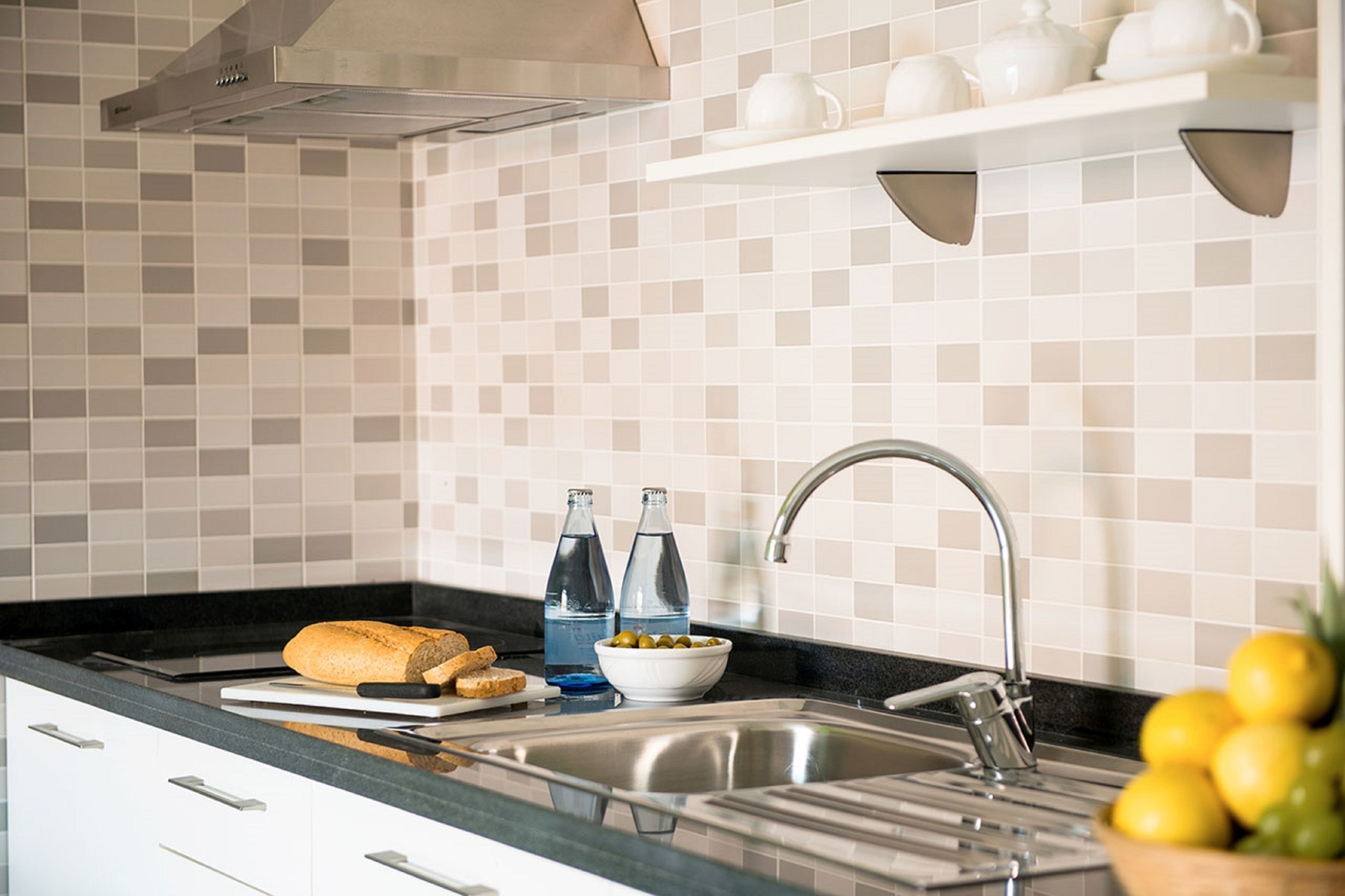 A modern kitchen with a stainless steel sink, a cutting board with bread, and a bowl of fruit.