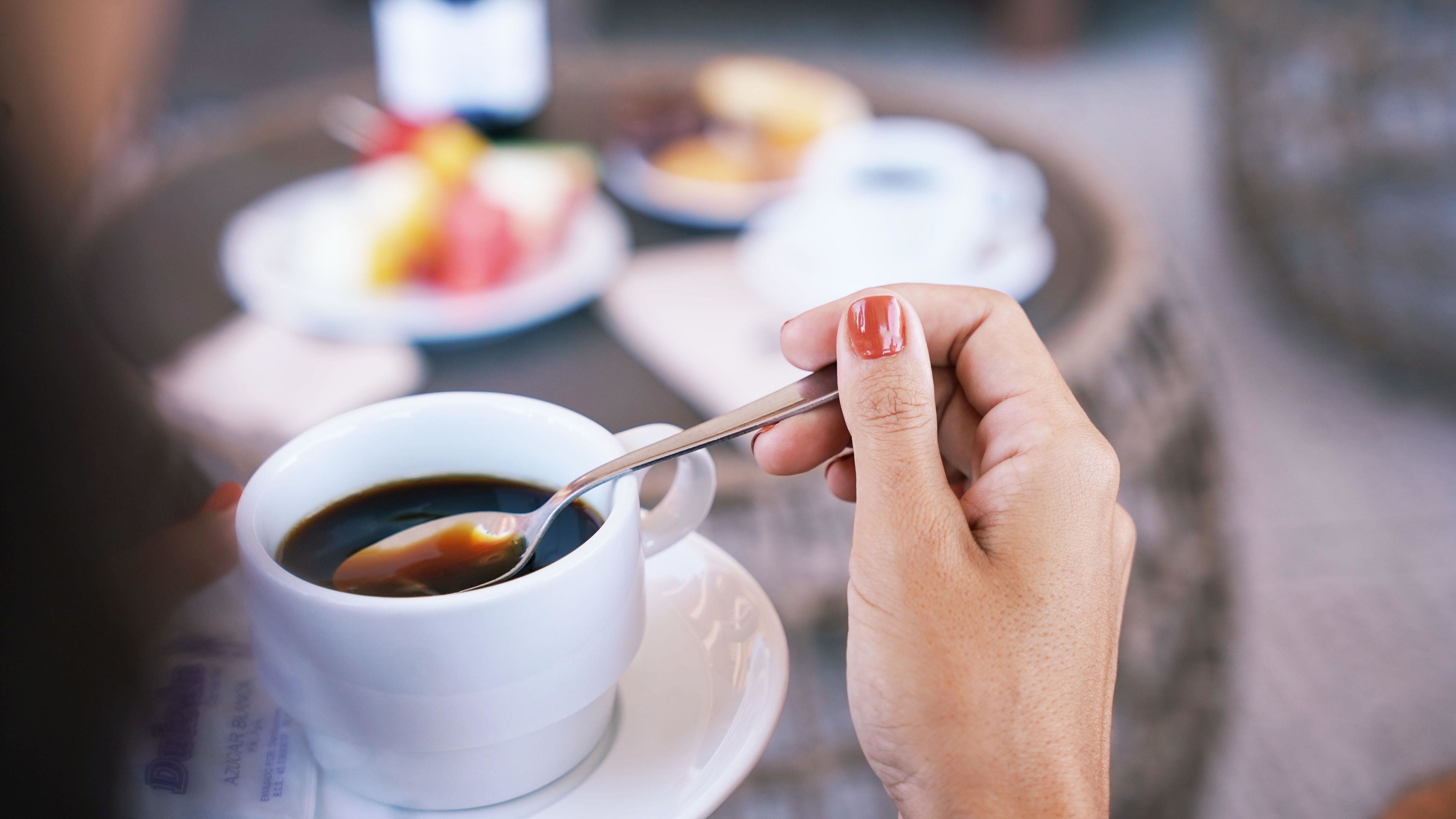 A hand holding a spoon over a cup of coffee, with a blurred background of a dining table with plates.
