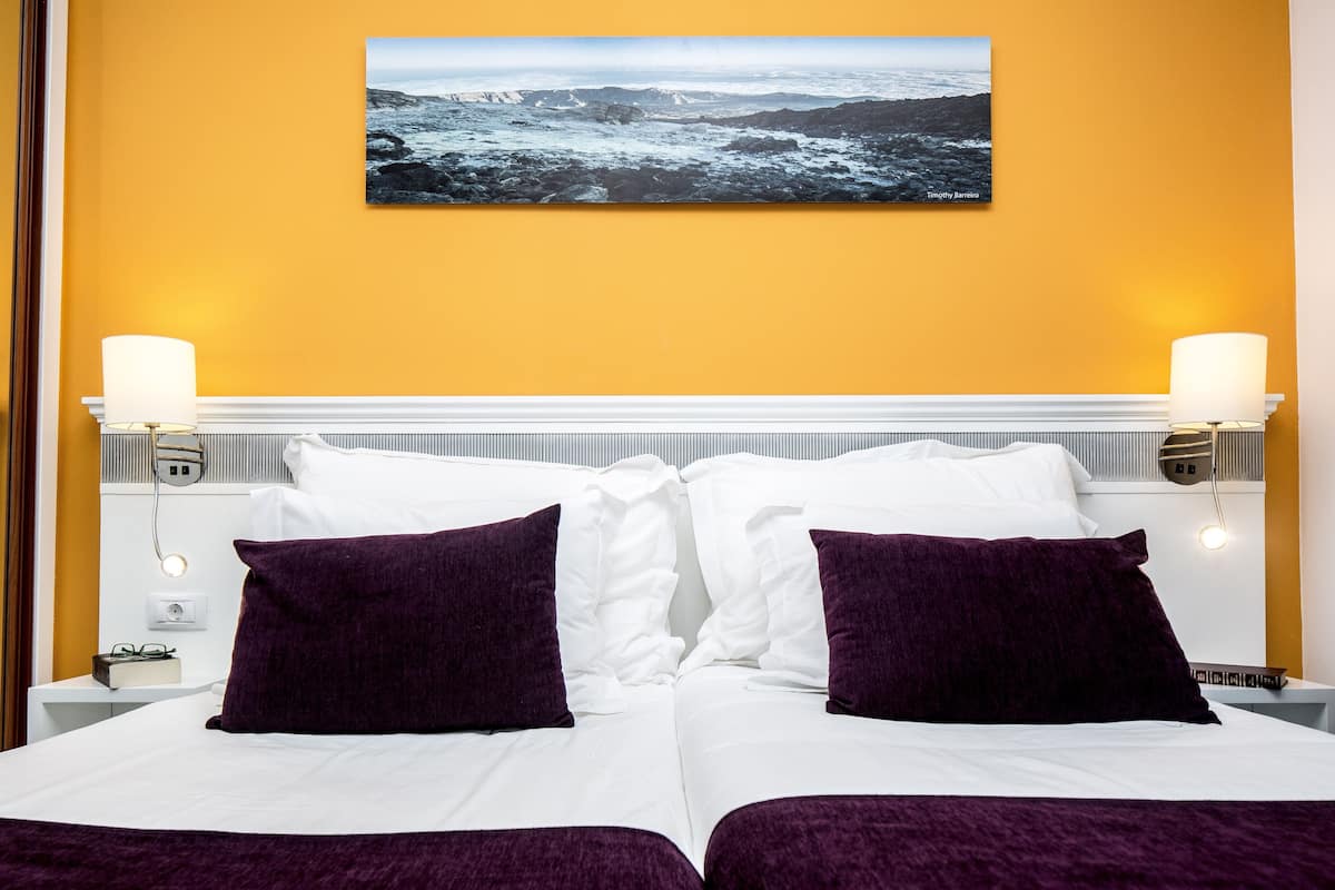 A hotel room with a bed, two purple pillows, a white headboard, and a picture of a rocky landscape on the wall.