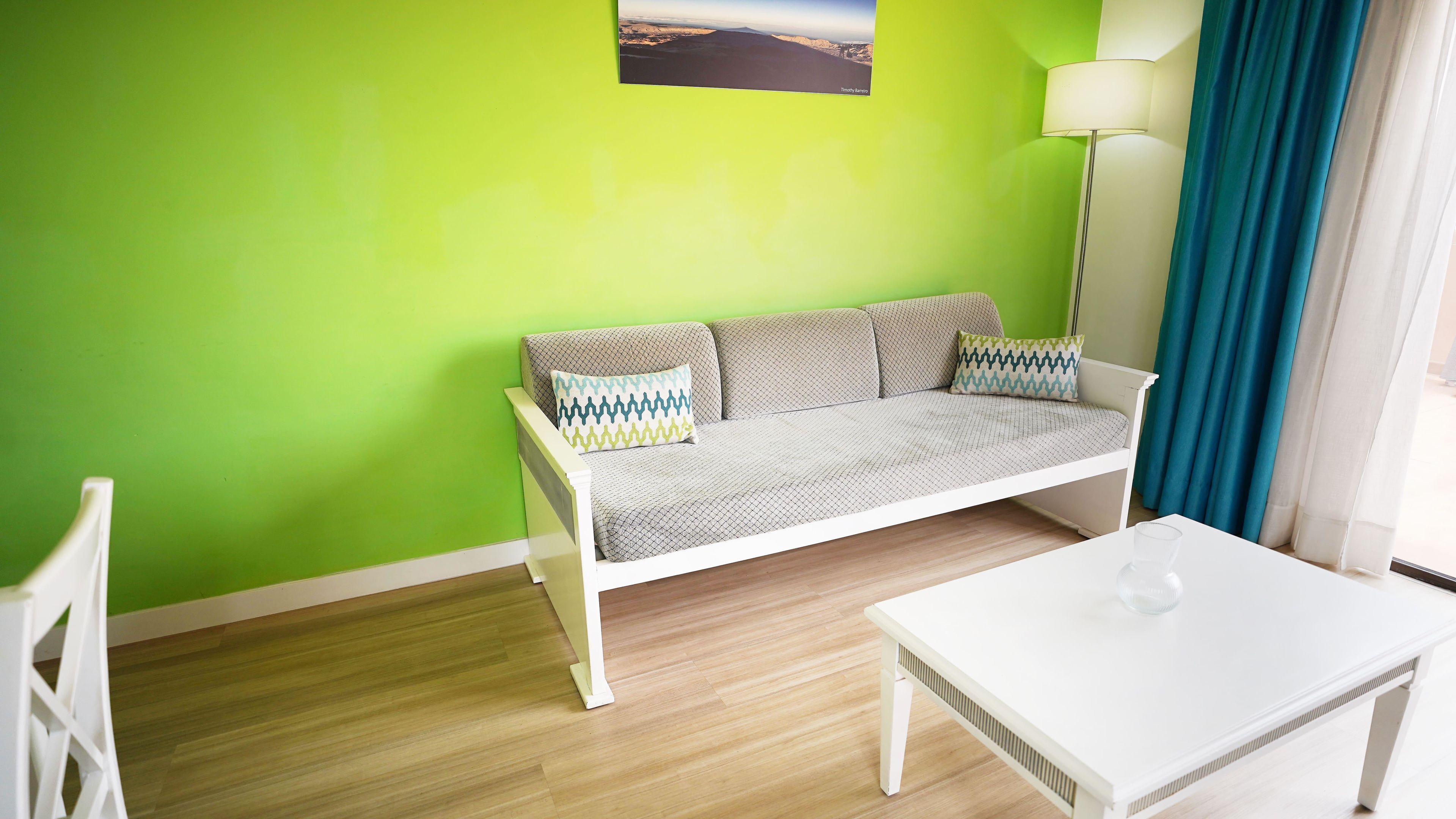 A room with a green wall, a white sofa with patterned cushions, a white table, and a floor lamp.
