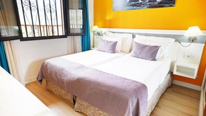 A double bed with a patterned bedspread, two pillows, a nightstand with a lamp, a power outlet, and a window with a curtain. - Coral Los Alisios (Arona)