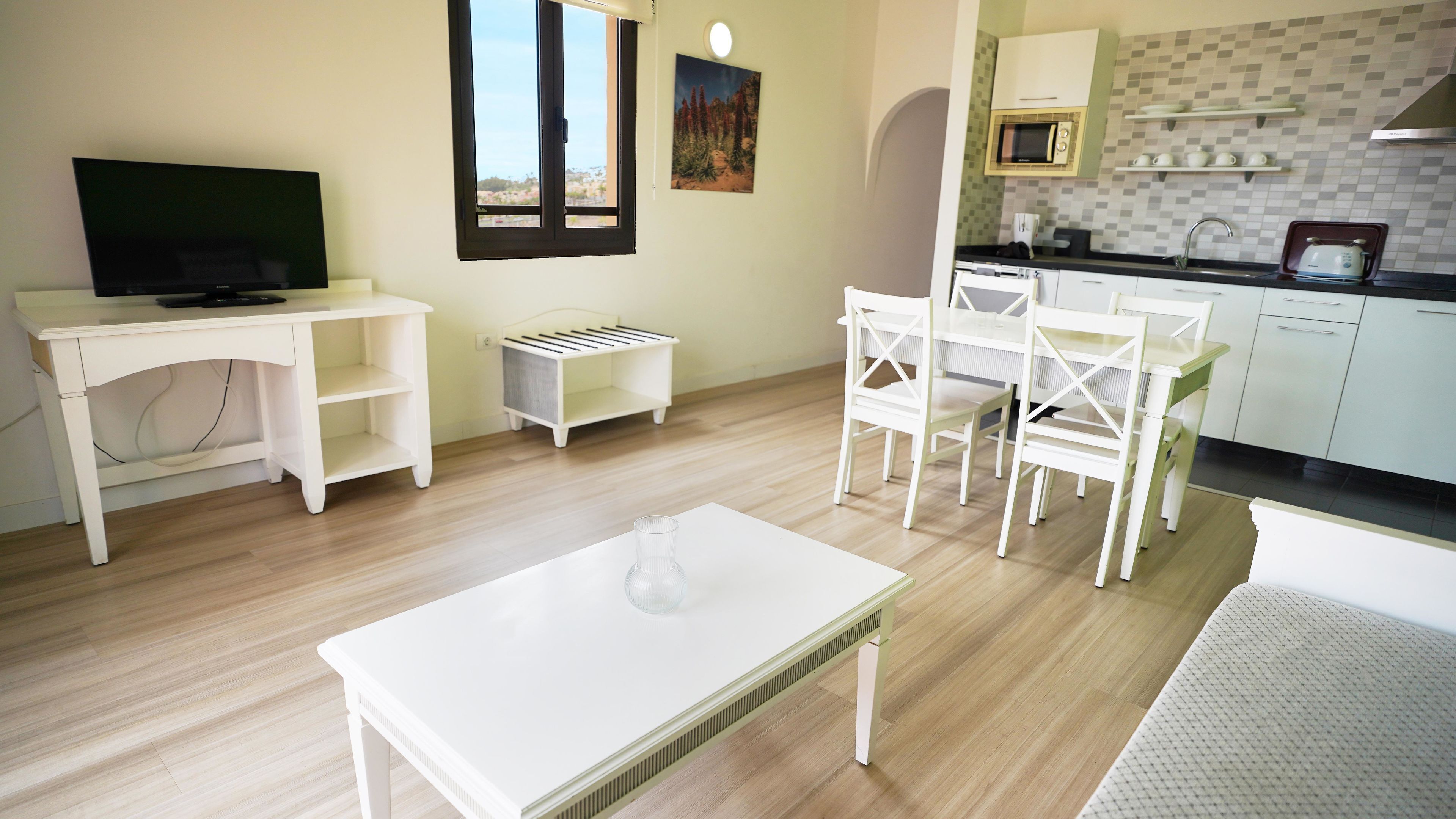 Una cocina moderna con armarios blancos, un microondas y un tostador. Hay un comedor con sillas blancas y una pequeña mesa. Un televisor está montado en la pared, y una ventana ofrece vistas al exterior.