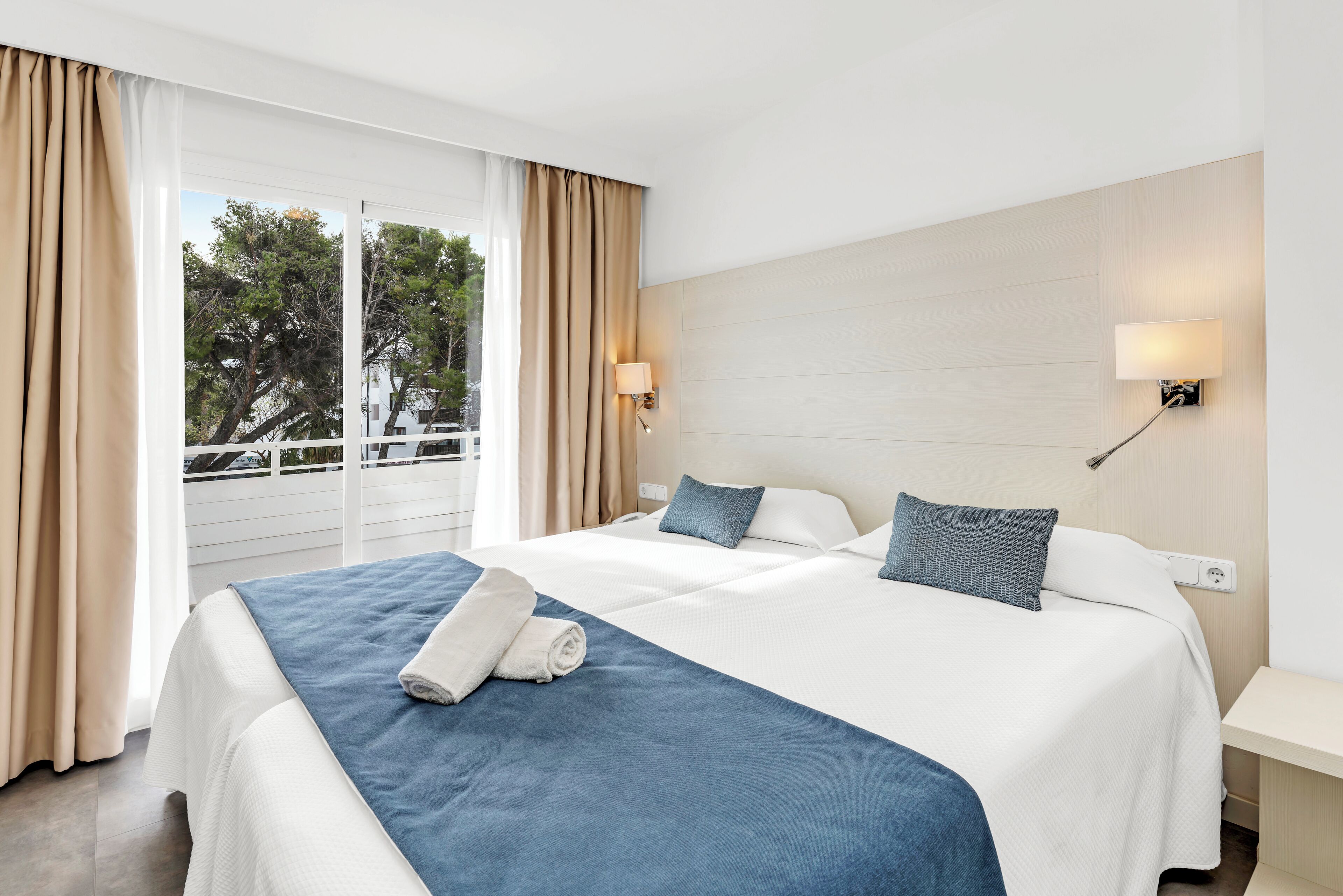 A hotel room with a bed, curtains, a nightstand, a wall-mounted lamp, and a view of trees through the window.