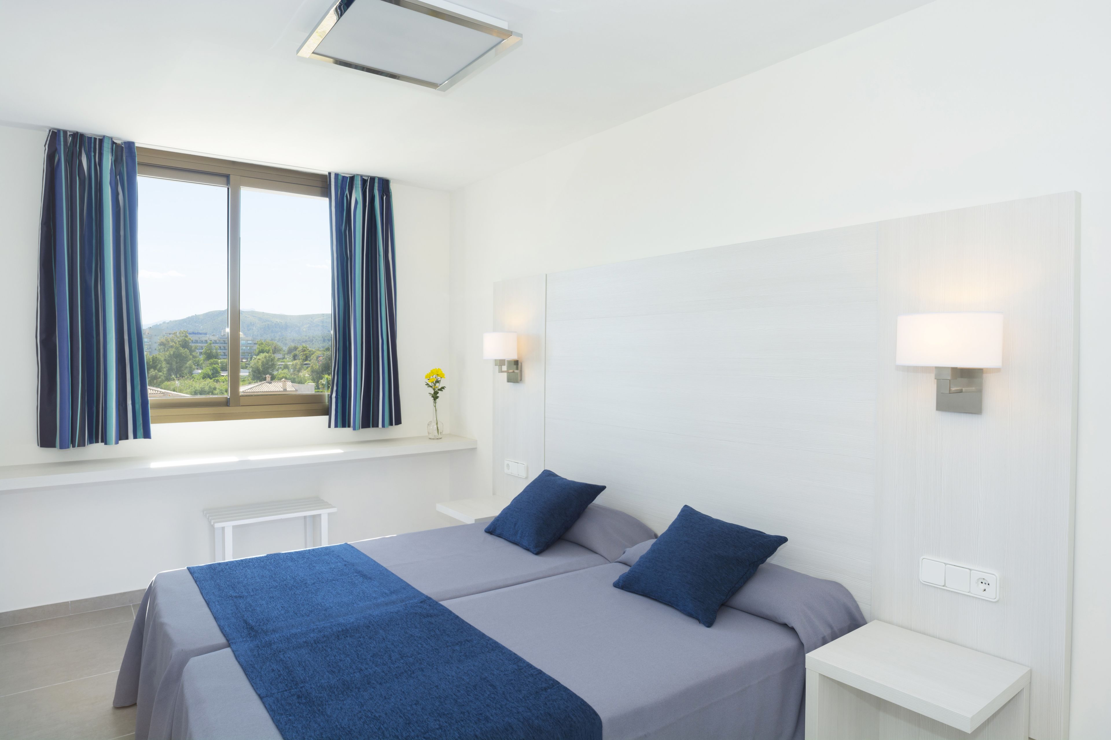 Een hotelkamer met twee bedden, een raam met gordijnen, een kleine tafel en een wandlamp.