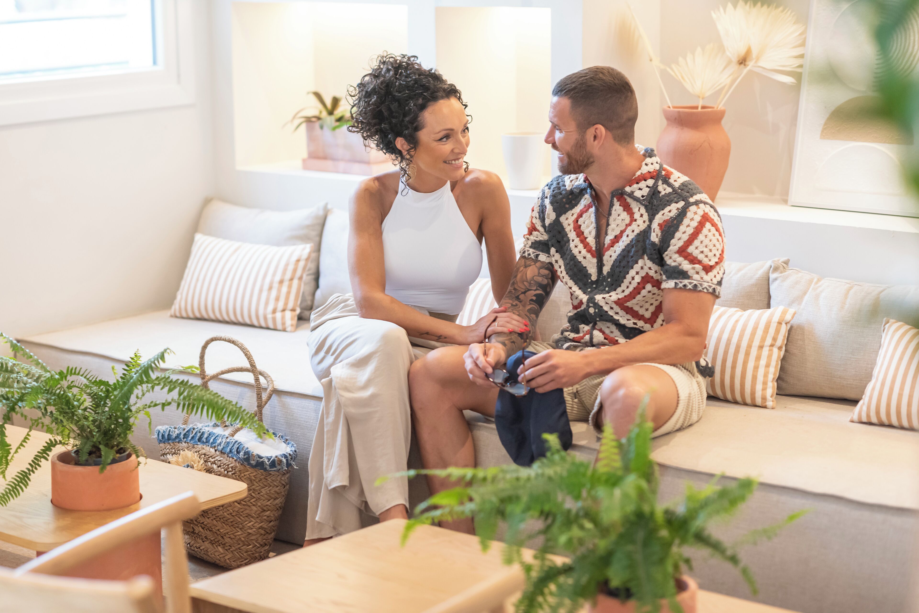 A couple sitting on a sofa in a well-lit room with a window, a plant, and a table with a basket.