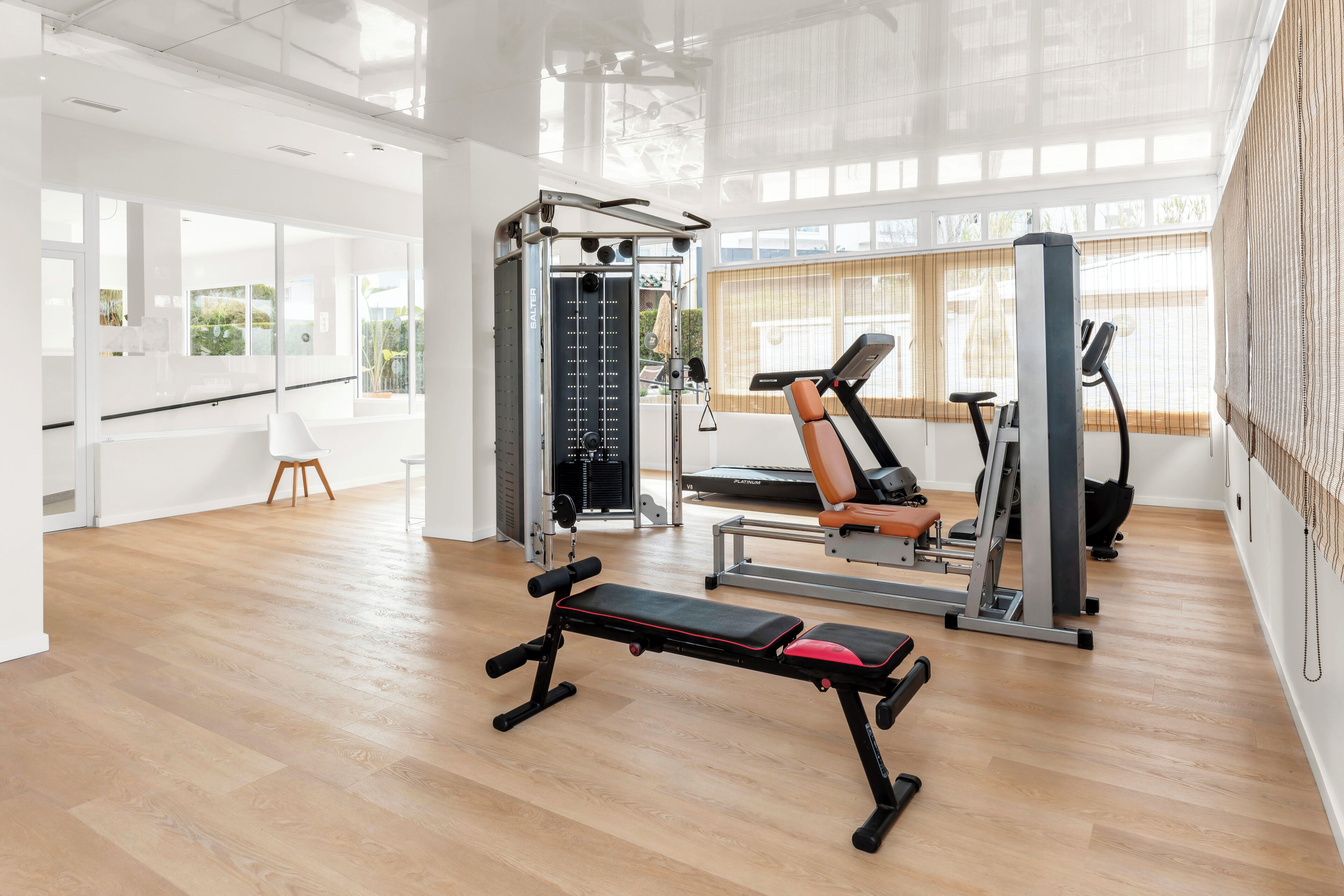 Un gimnasio con equipamiento de ejercicio, una banca y ventanas amplias.