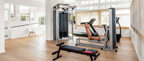 Un gimnasio con equipamiento de ejercicio, una banca y ventanas amplias.