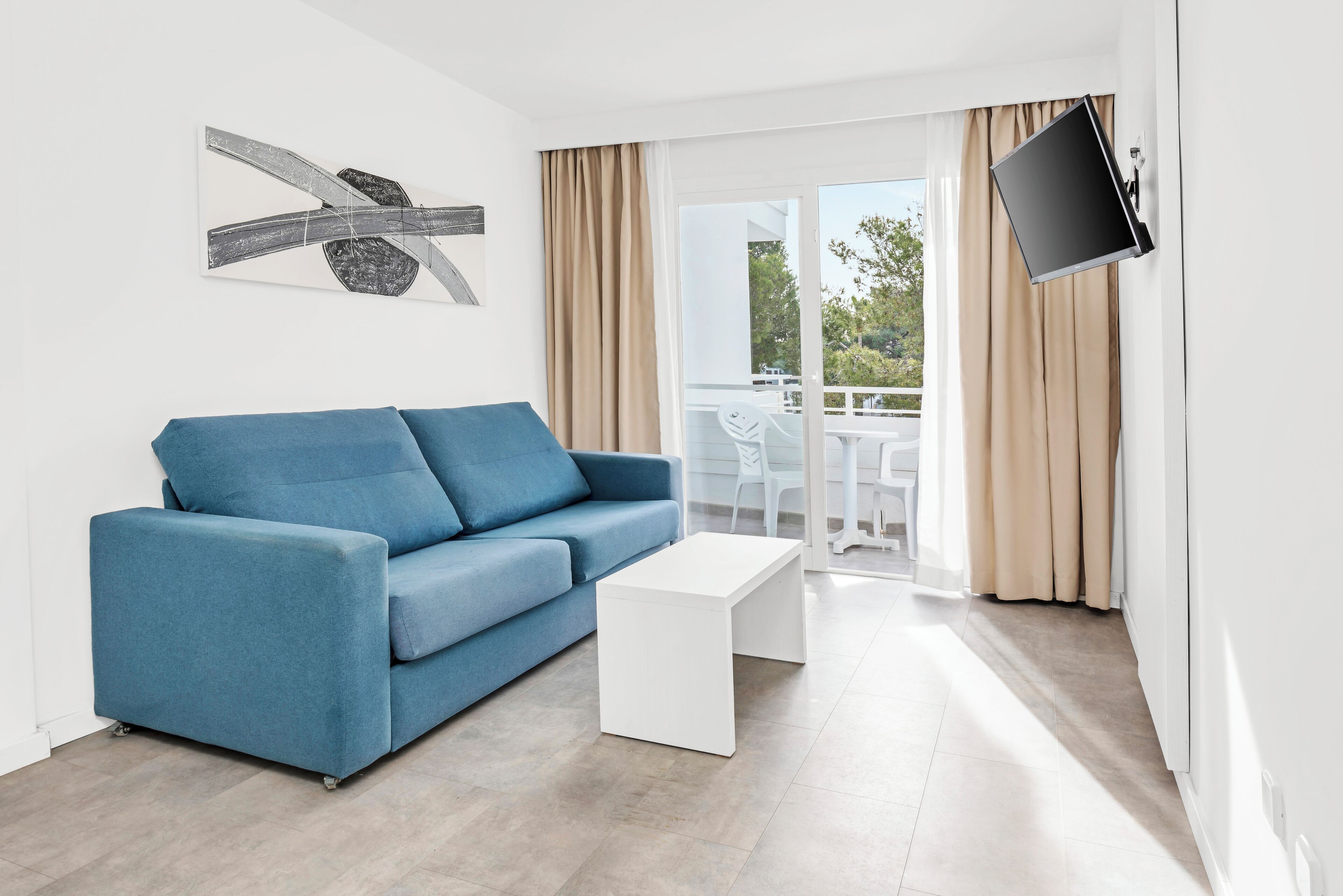 A modern living room with a blue sofa, a white coffee table, a flat-screen TV mounted on the wall, and a balcony with a table and chairs.