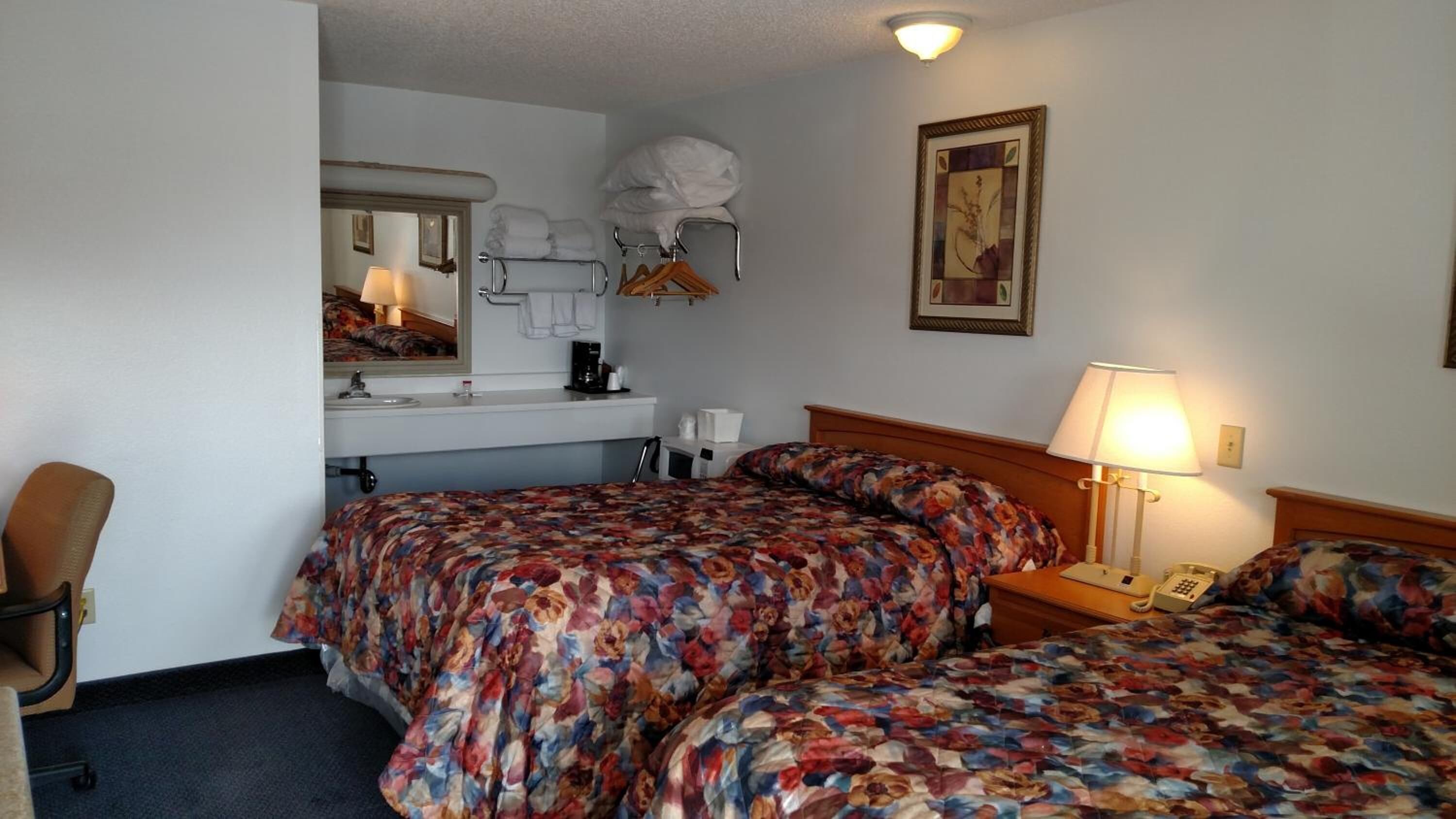 A hotel room with two beds, a desk with a lamp, a chair, a bathroom mirror, and a framed picture on the wall.