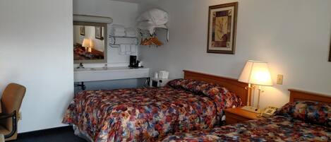A hotel room with two beds, a desk with a lamp, a chair, a bathroom mirror, and a framed picture on the wall.