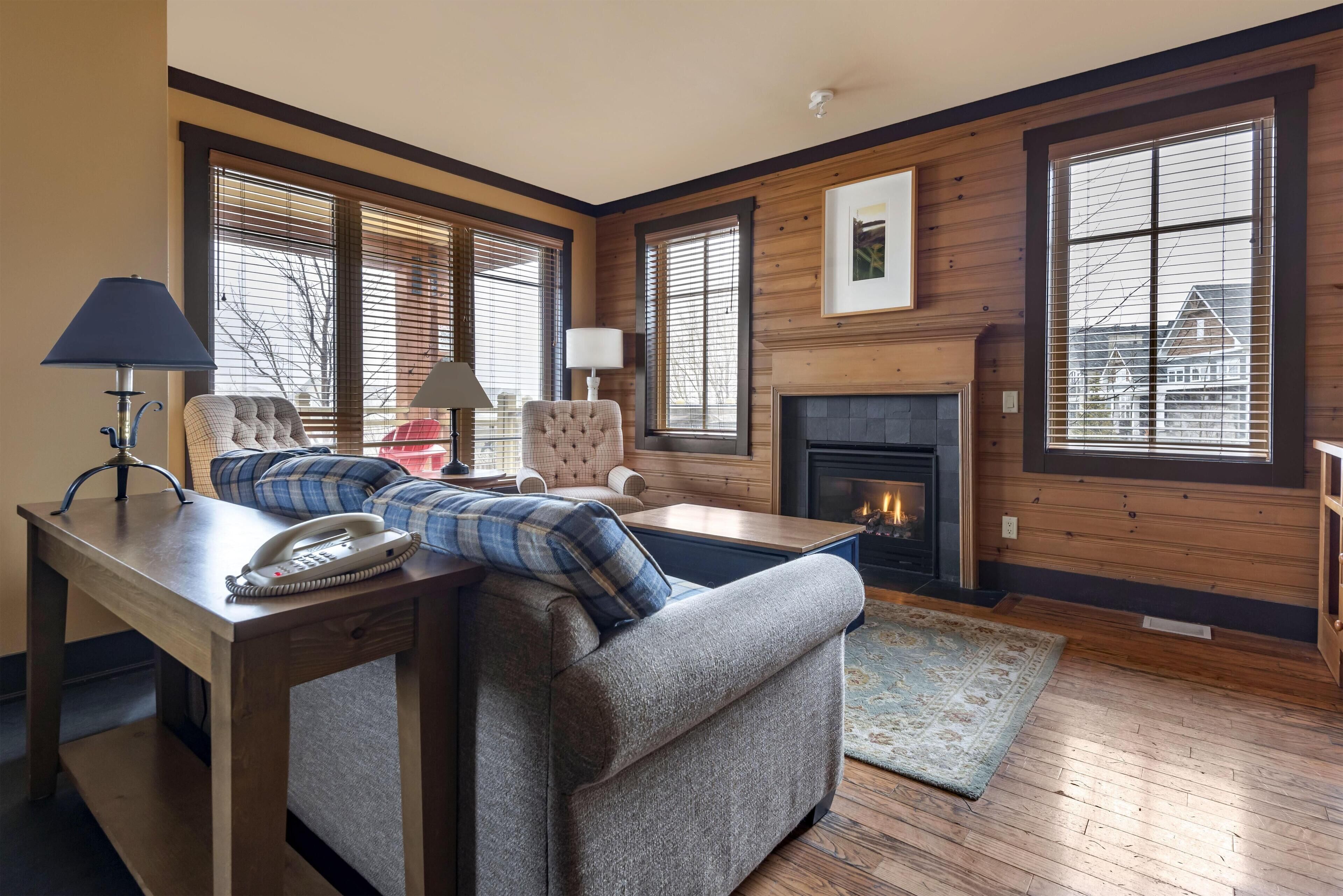 A cozy living room with a fireplace, a sofa, a chair, and a table with a lamp and a phone.