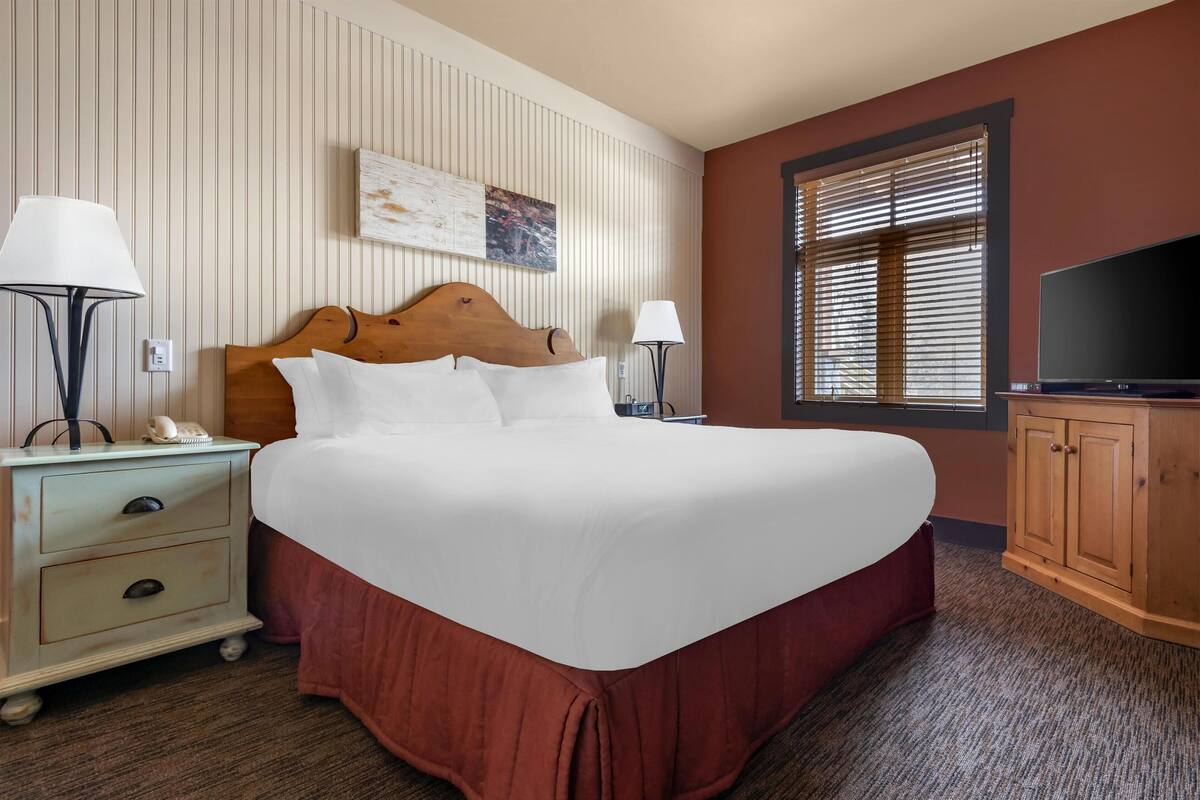 Una habitación de hotel con una cama grande, una mesita de noche, una lámpara, un televisor y una ventana con persianas.