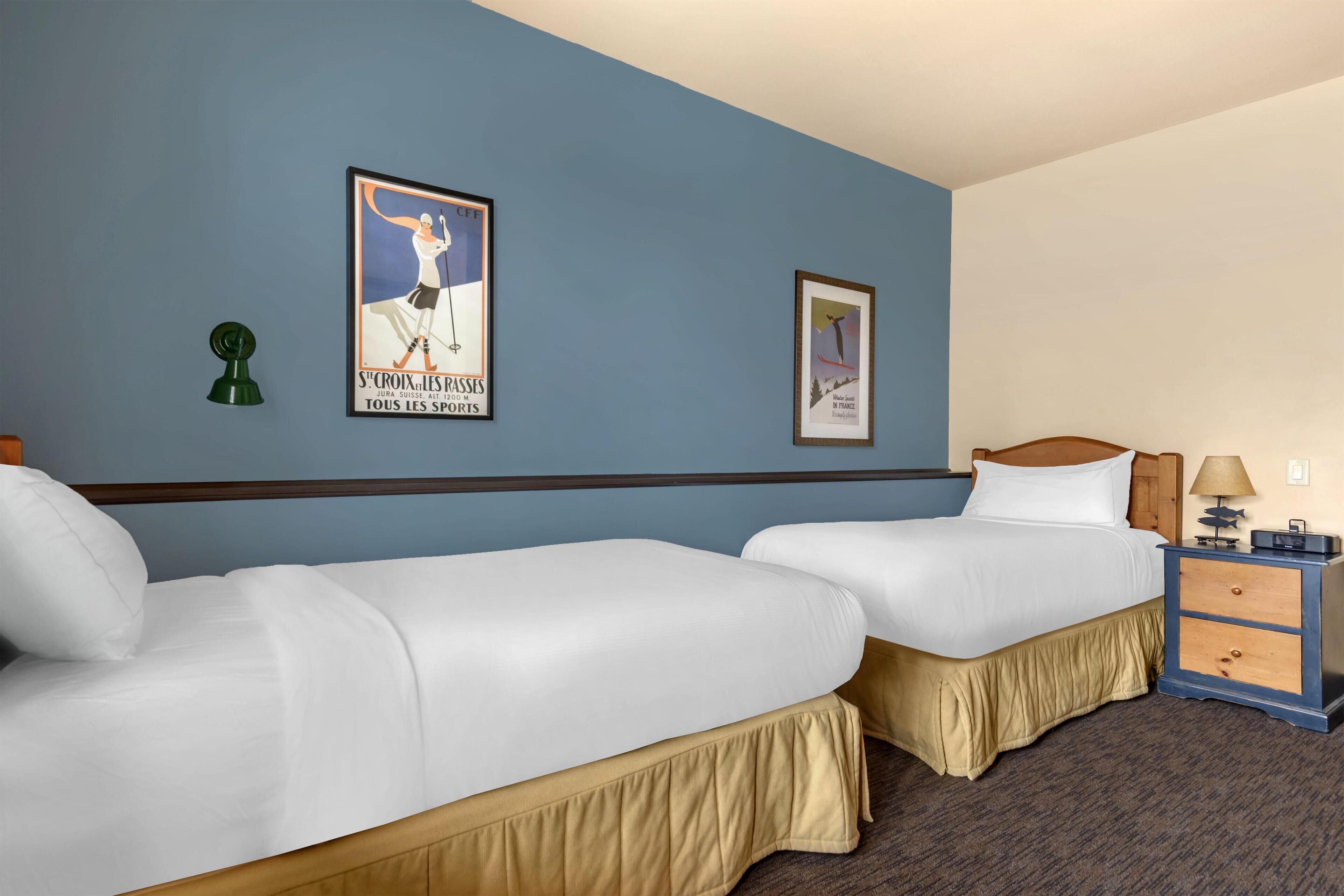 A hotel room with two single beds, a nightstand with a lamp, and two wall-mounted posters.