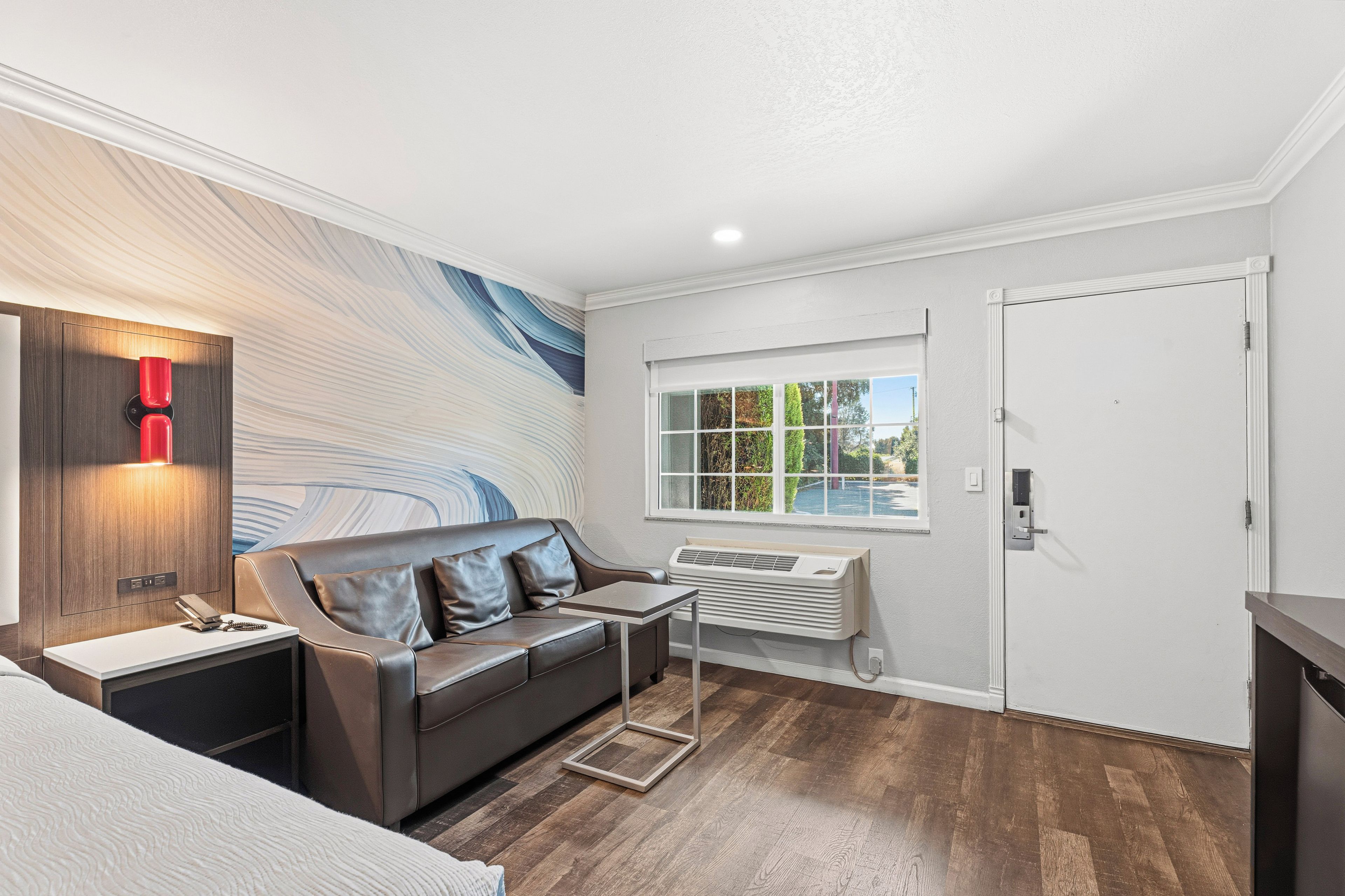 A modern room with a sofa, a desk, a wall-mounted heater, and a window with a view of greenery.