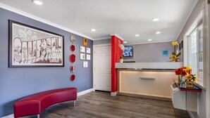 Un área de recepción moderna con un banco rojo, un cartel de bienvenida enmarcado y un televisor montado en la pared.