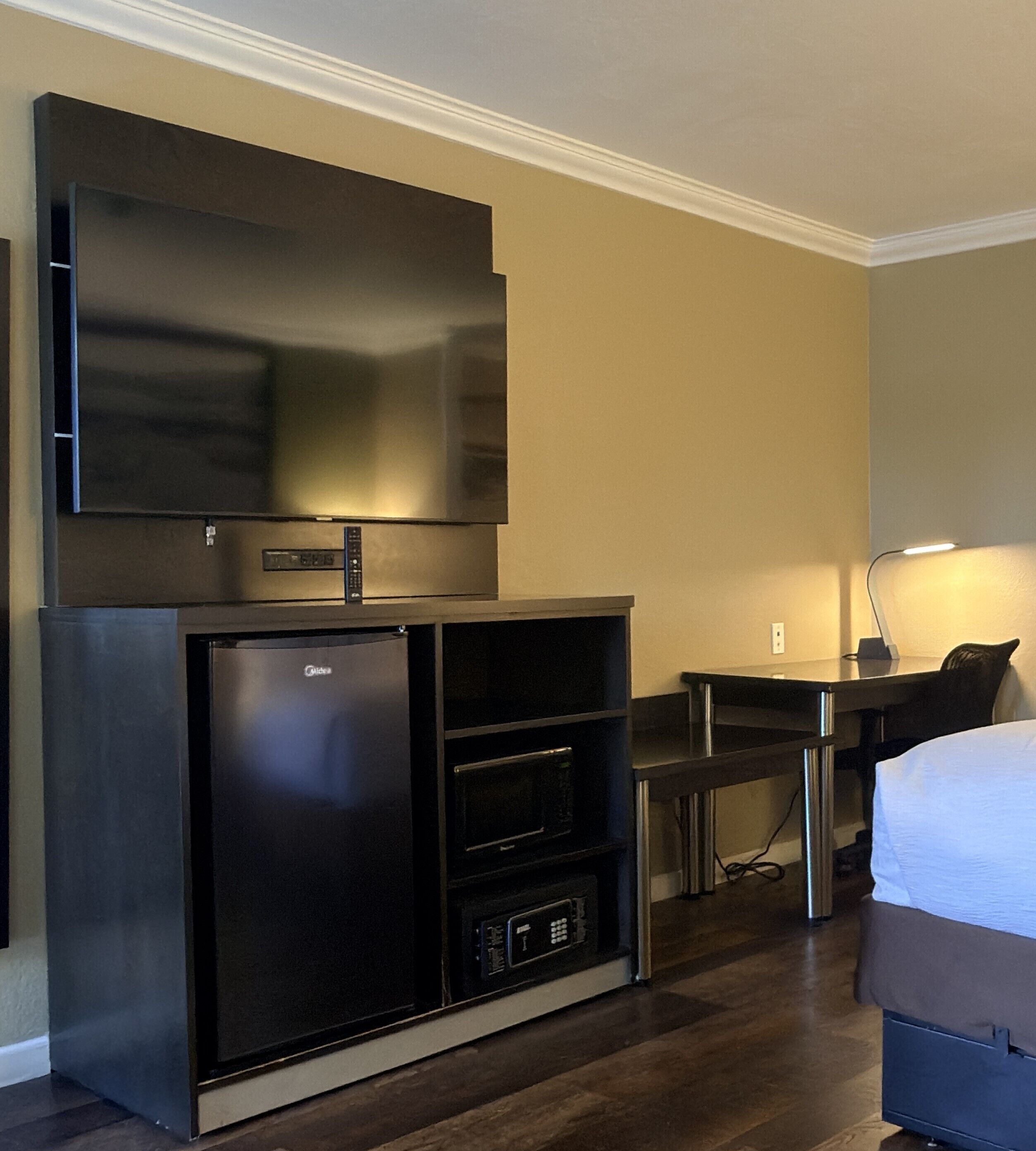 A compact hotel room with a kitchenette, a flat-screen TV, a bed, and a desk with a lamp.