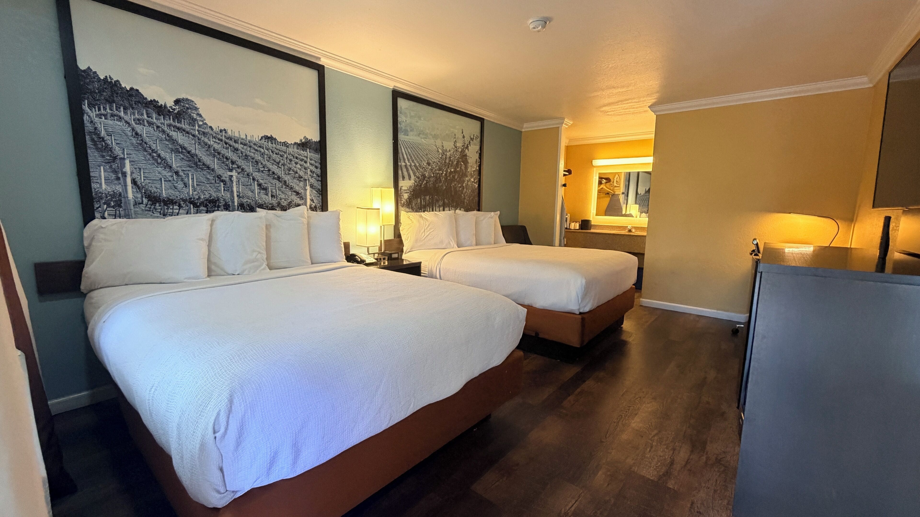 Habitación de hotel con dos camas, un cuadro grande en la pared, un escritorio con lámpara y una pequeña zona de kitchenette.