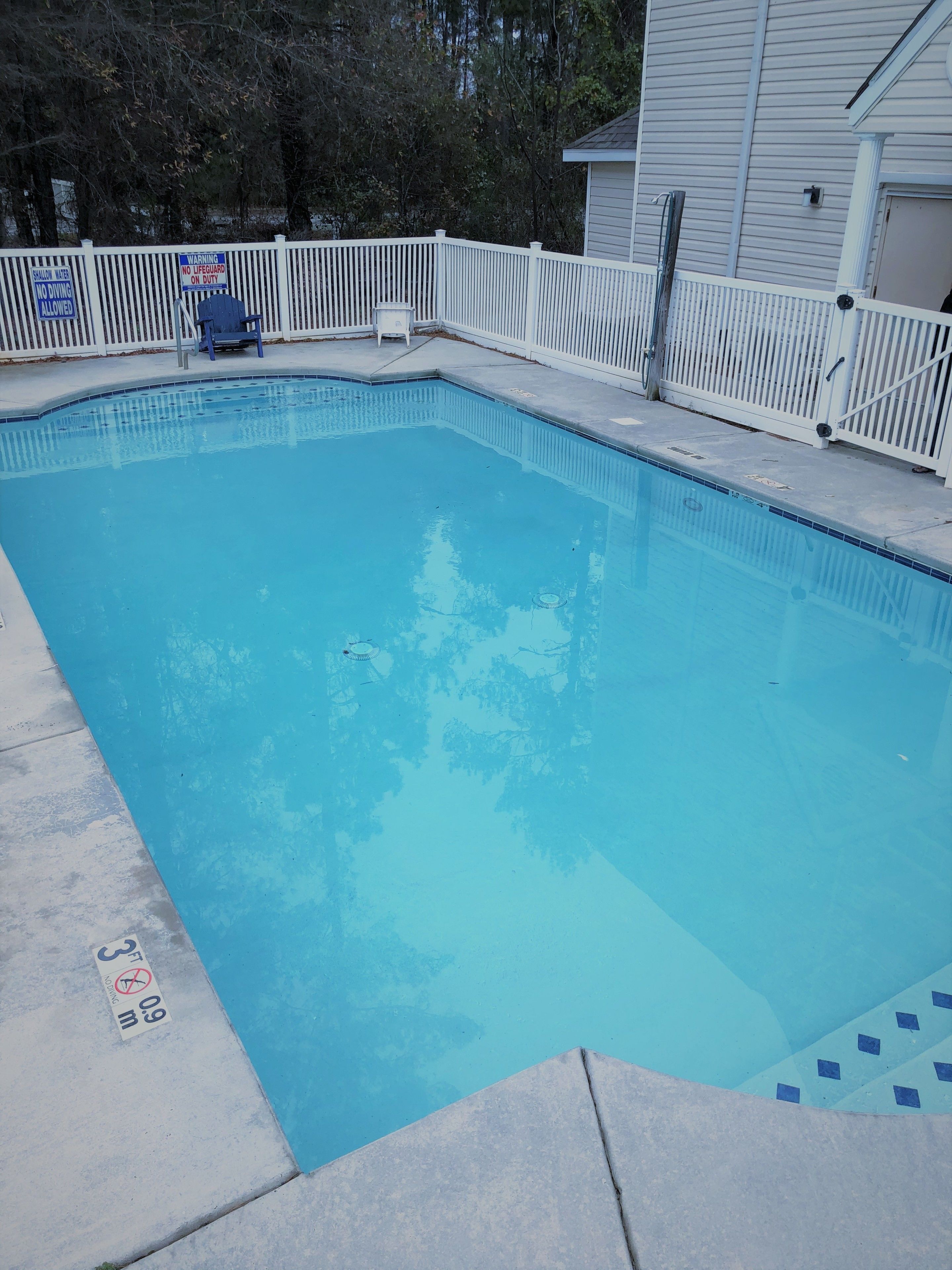 Une piscine rectangulaire entourée d’une clôture blanche, avec un panneau d’avertissement indiquant une faible profondeur et un panneau indiquant la présence d’un sauveteur.