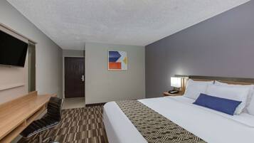 Quarto de hotel com cama, escrivaninha, cadeira, televisão e porta.