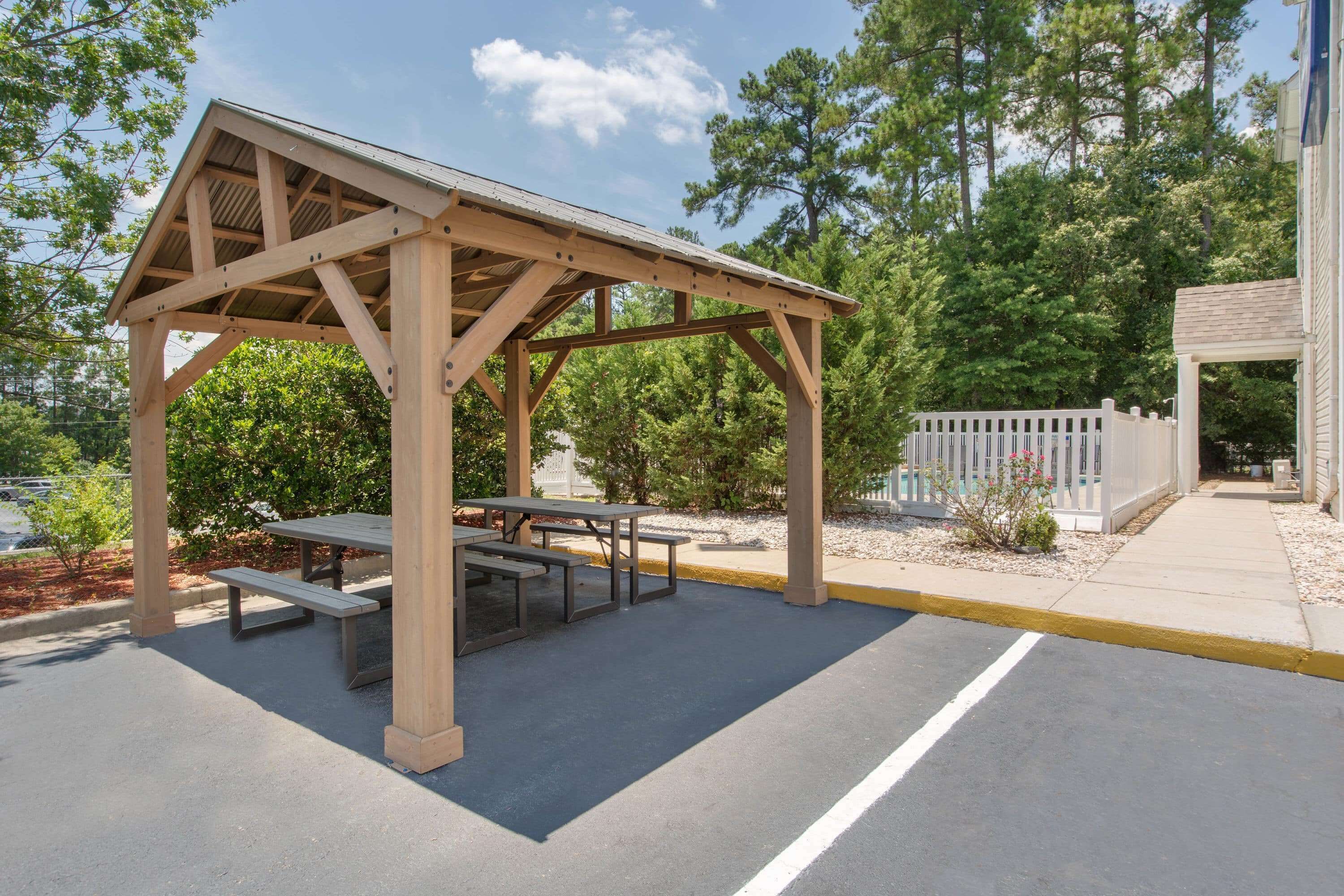 A wooden pavilion with picnic tables, surrounded by trees and a fenced area.