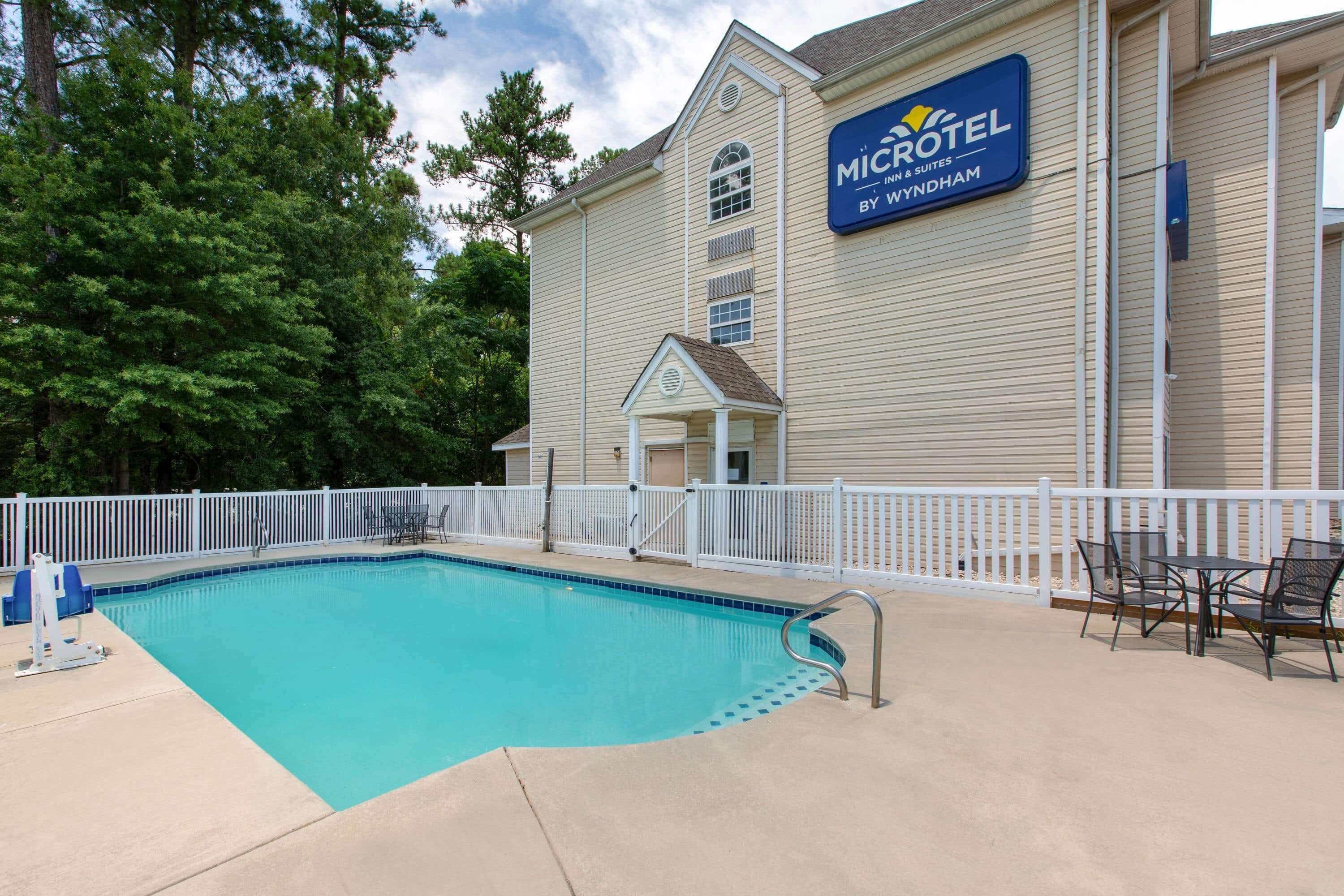 Une piscine entourée d’une clôture blanche, un espace patio avec des chaises, et un bâtiment sur lequel on peut lire l’inscription « MICROTEL INN & SUITES BY WYNDHAM ».