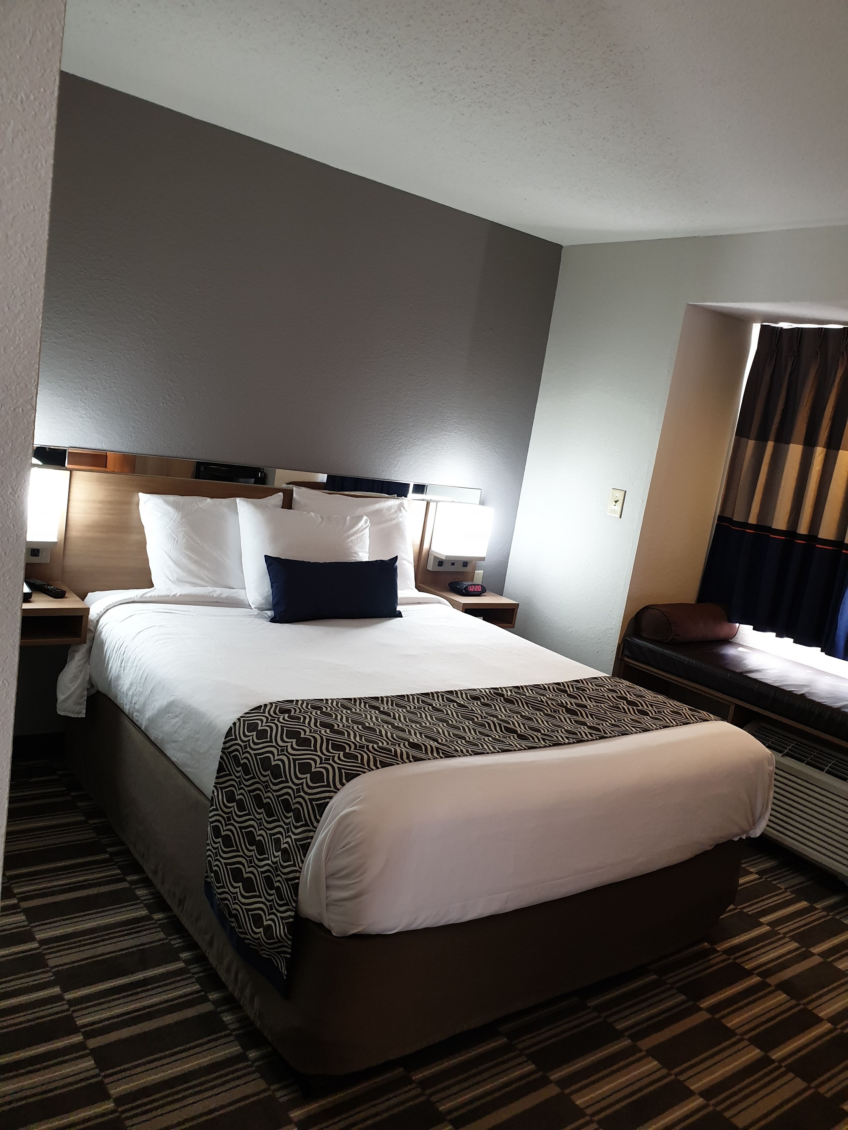A hotel room with a large bed, bedside tables, a nightstand, a window with curtains, and a patterned carpet.