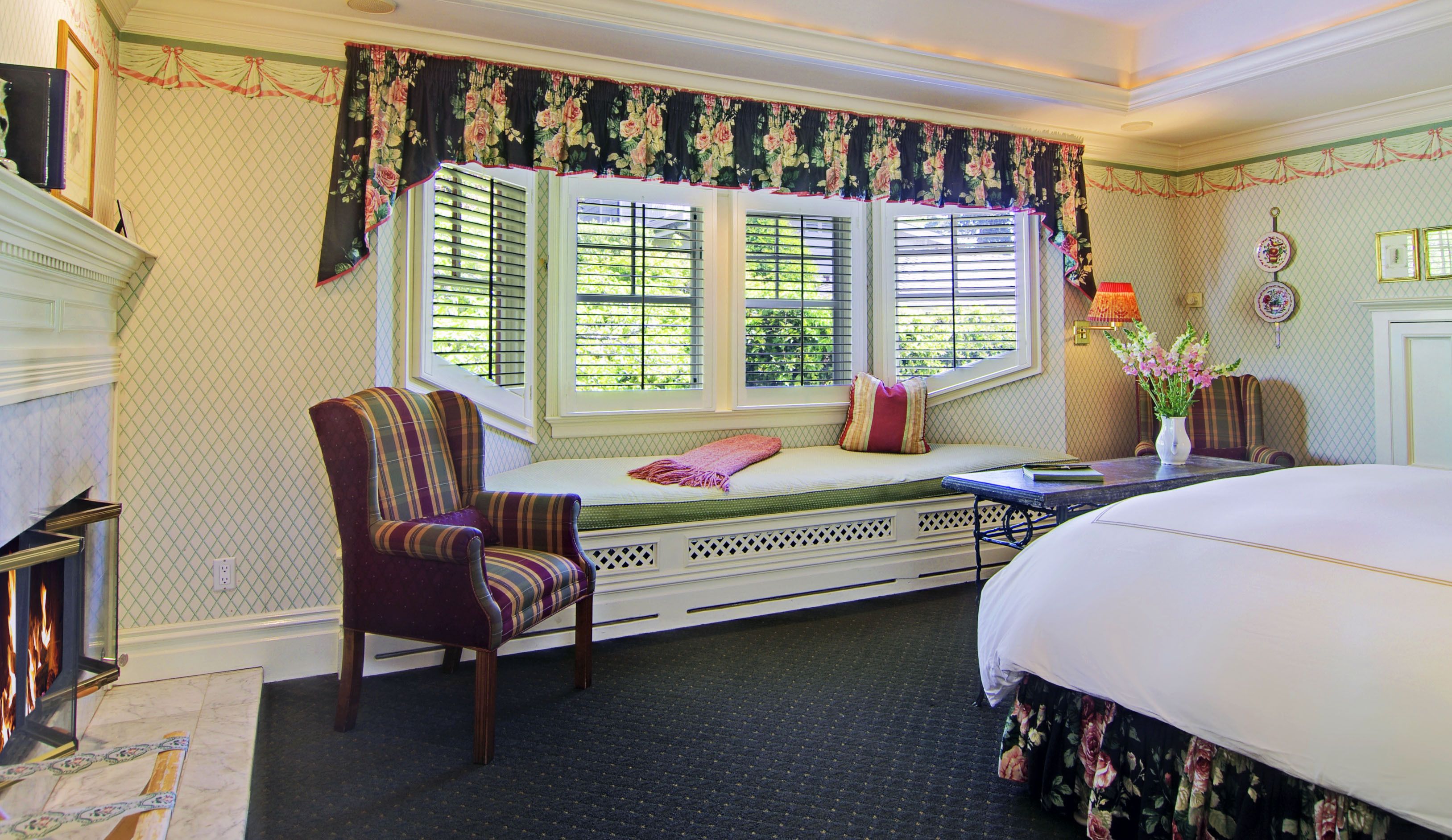 A bedroom with a large bed, a window seat with a chair, and a fireplace.