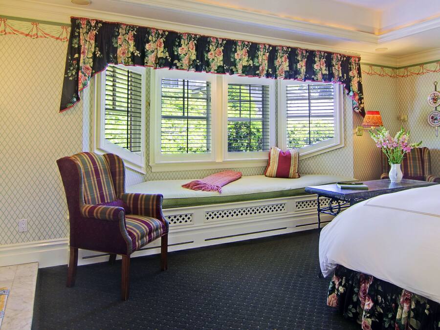 A bedroom with a large bed, a window seat with a chair, and a fireplace.